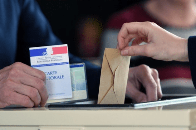 Une main tenant une enveloppe marron est en train de la déposer dans une urne électorale, tandis qu'une autre main présente une carte électorale française.